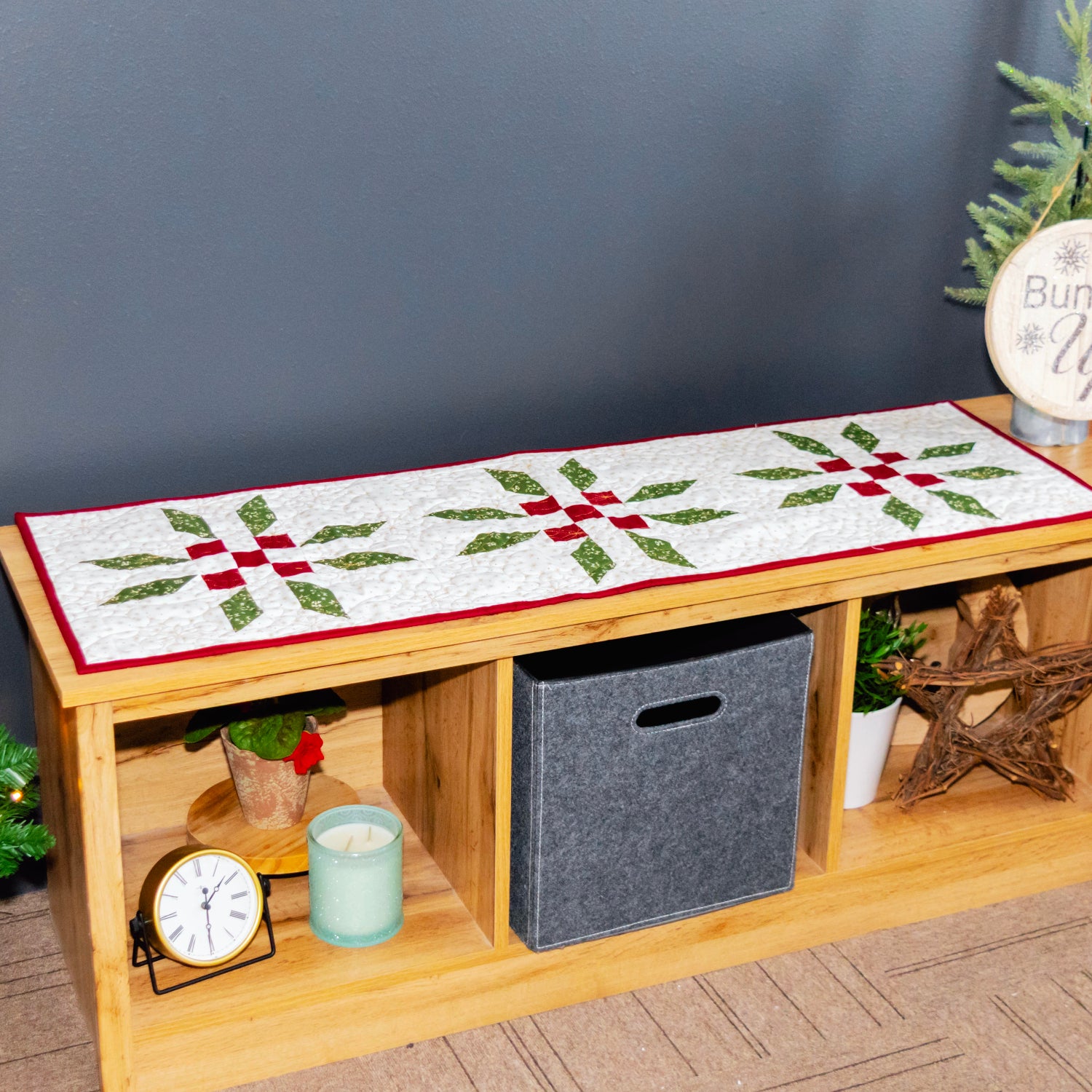 A wooden bench with open storage compartments, decorated with a white quilted holiday table runner featuring green pine branches and red bows. The bench is styled in a cozy room setting with a Christmas tree, a gray wall, and a ladder shelf in the background.