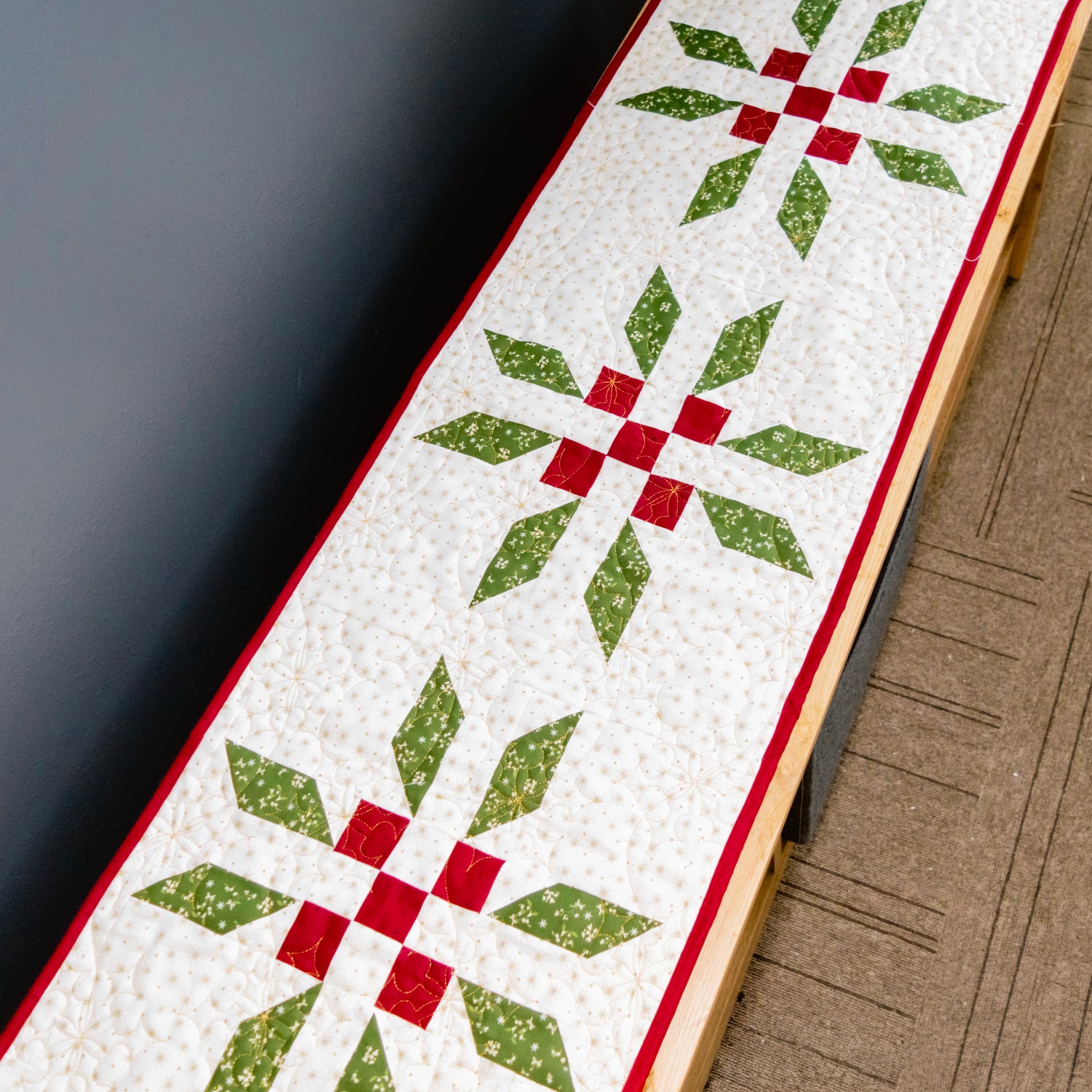 A close-up of a quilted table runner with a repeating green and red starburst pattern on a white background, shown resting on a carpeted floor next to a dark wall.
