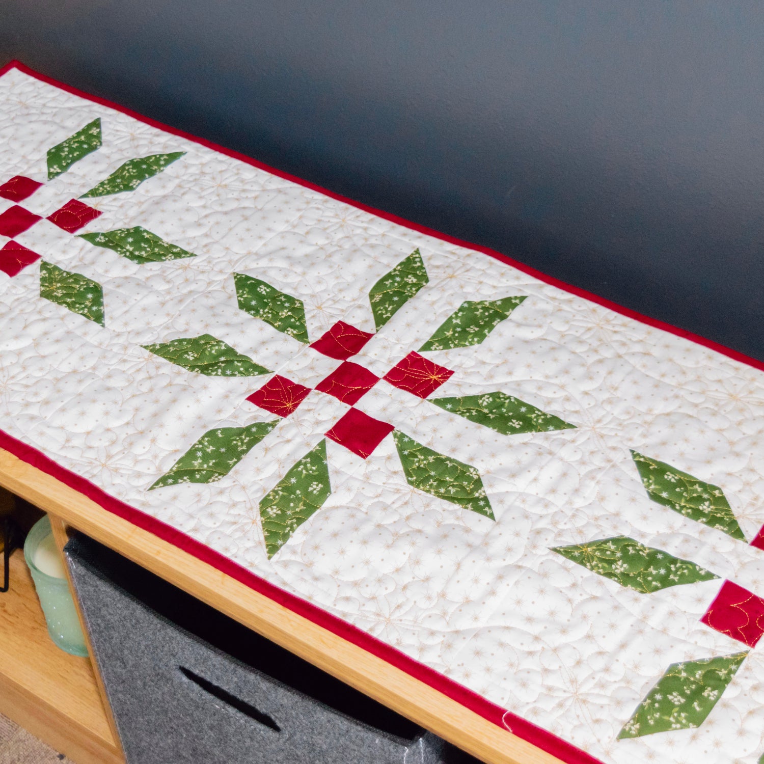 A quilted table runner with a white background, green starburst motifs, and red center squares, draped over a wooden bench.