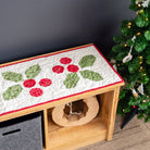 Decorative table runner with holly design on a wooden bench next to a Christmas tree.