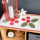 Quilted table runner with holly design on a wooden table, surrounded by Christmas decor.