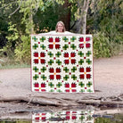 A woman stands outdoors on a dirt path holding up a large quilted blanket with a repeating red and green geometric pattern on a cream background. The scene is reflected in a body of water below, with trees and foliage behind her.