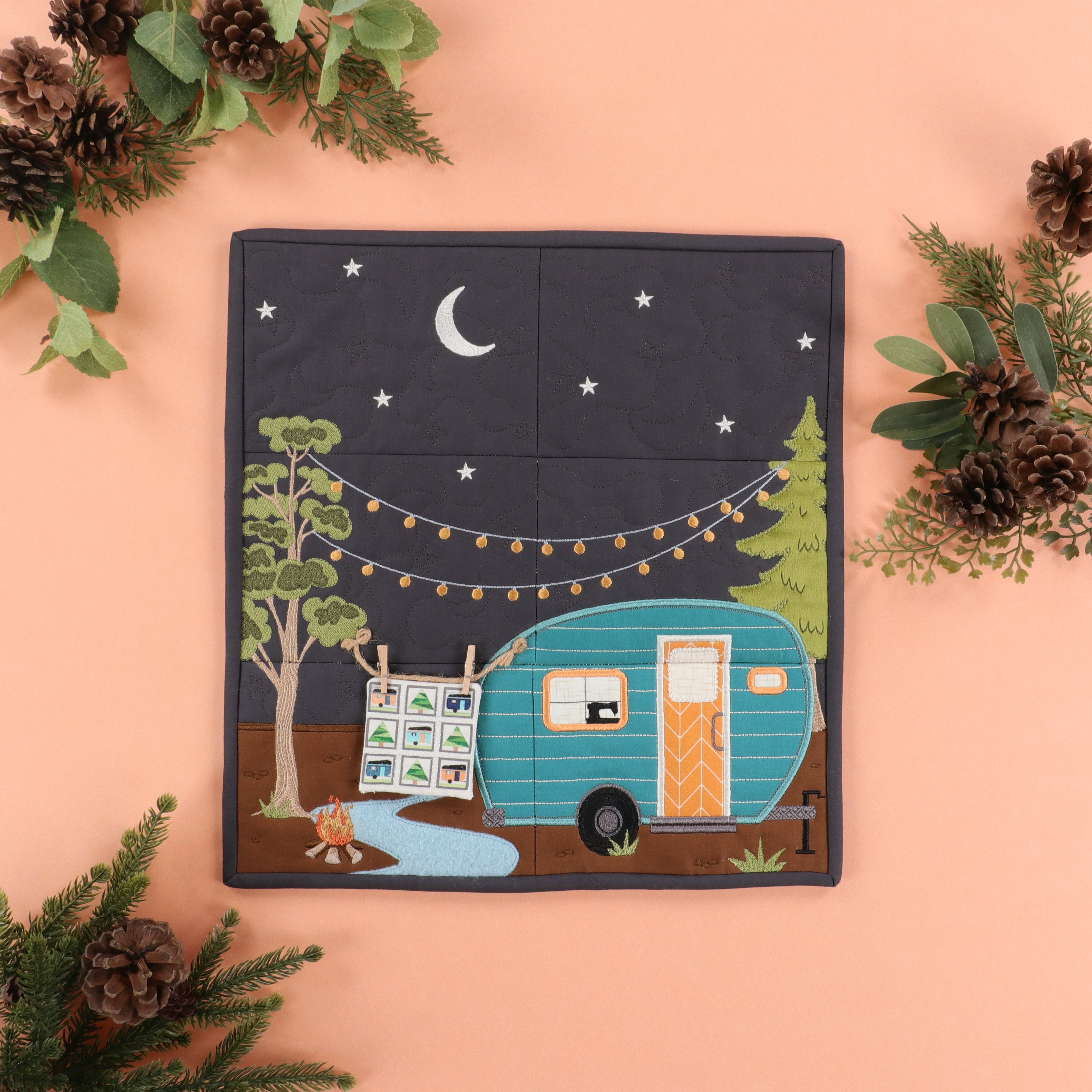 Patchwork quilt with a camper design surrounded by pinecones and greenery on a peach background