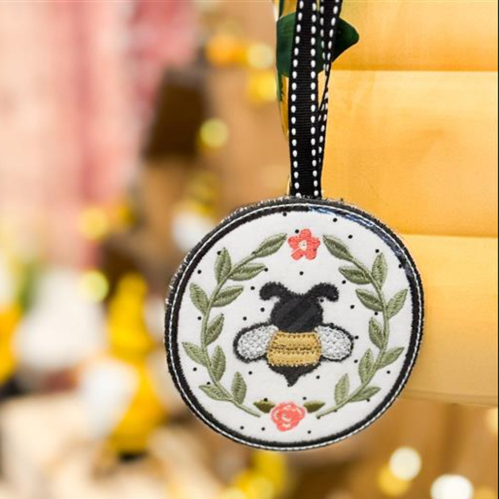 A circular honeybee-themed bag tag with embroidery detailing, held against a blurred background.