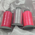 Three spools of thread on gray plastic cores, with two spools wrapped in bright pink thread and one wrapped in clear thread, arranged on crumpled white tissue paper.