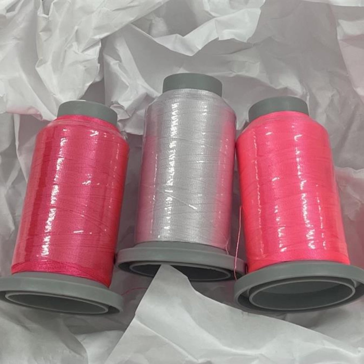 Three spools of thread on gray plastic cores, with two spools wrapped in bright pink thread and one wrapped in clear thread, arranged on crumpled white tissue paper.