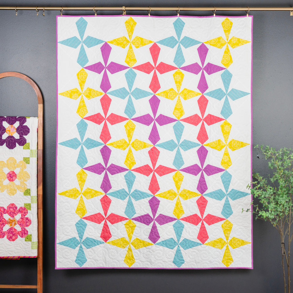 Colorful quilt with geometric pattern on a gray wall, next to a smaller floral quilt on a wooden ladder.