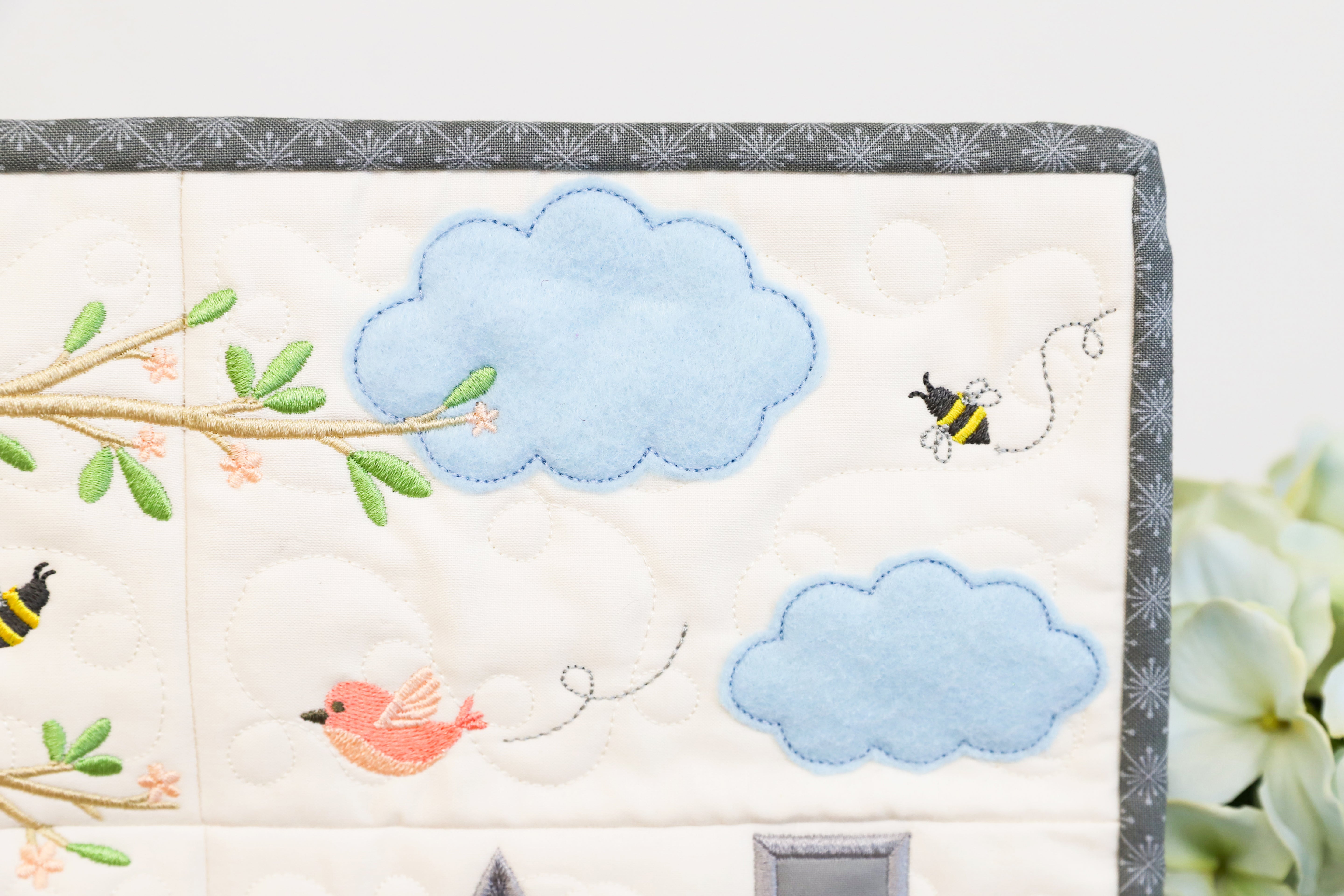Embroidered panel with blue clouds, a branch with leaves, and a bee on a white background.