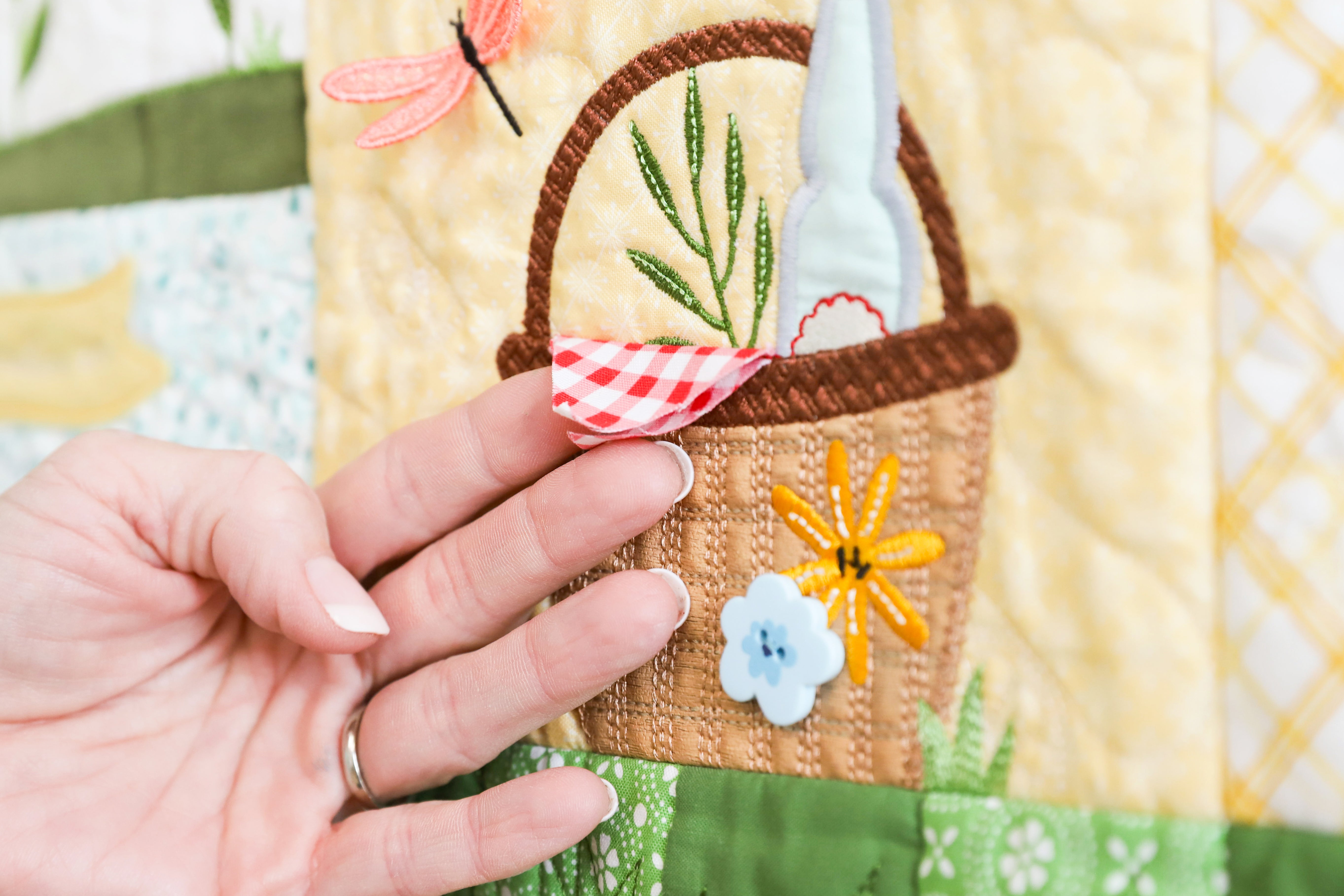 Hand holding a small embroidered basket with flowers on a patchwork background