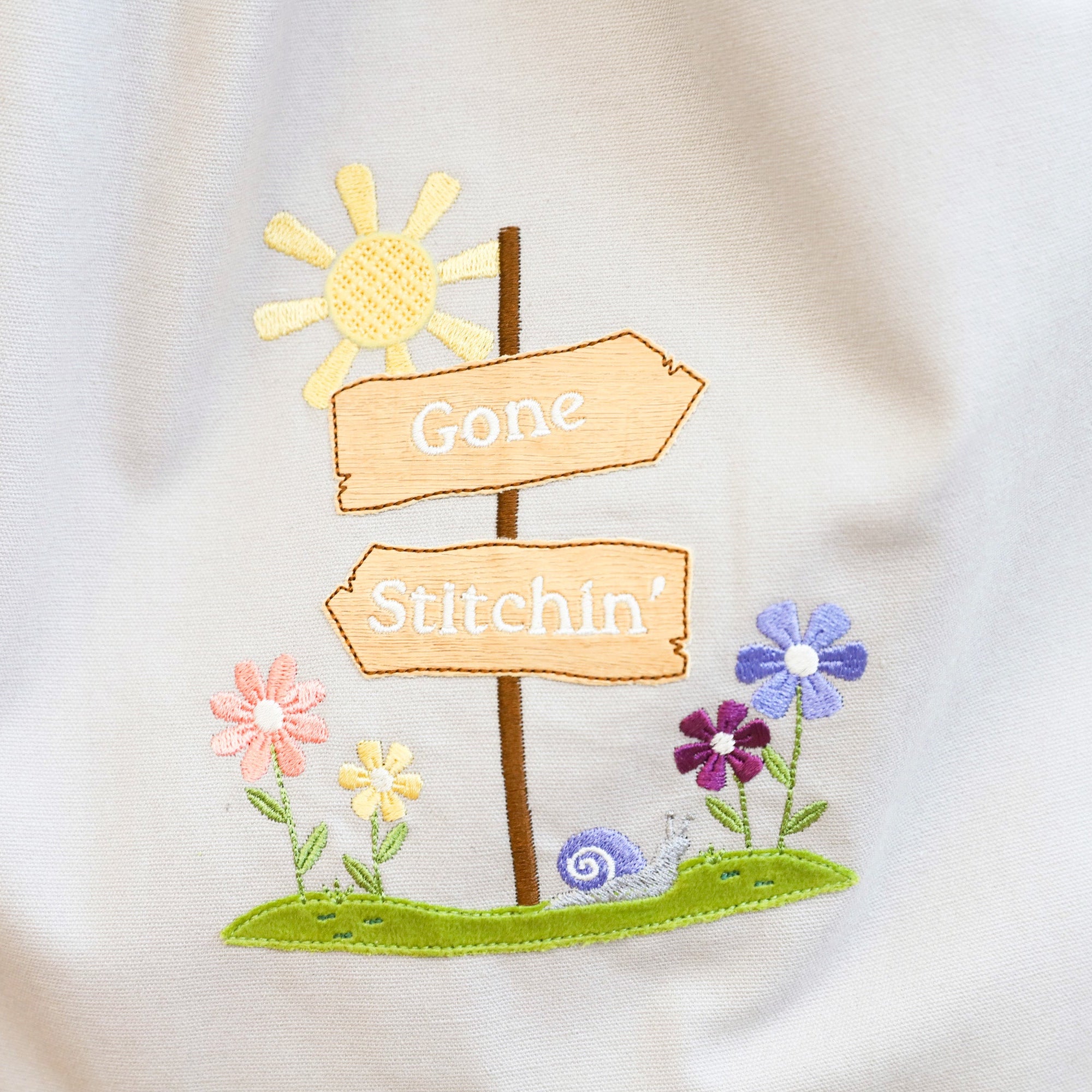 Embroidered design on a white fabric with signs and flowers, featuring the text 'Gone Stitchin'.
