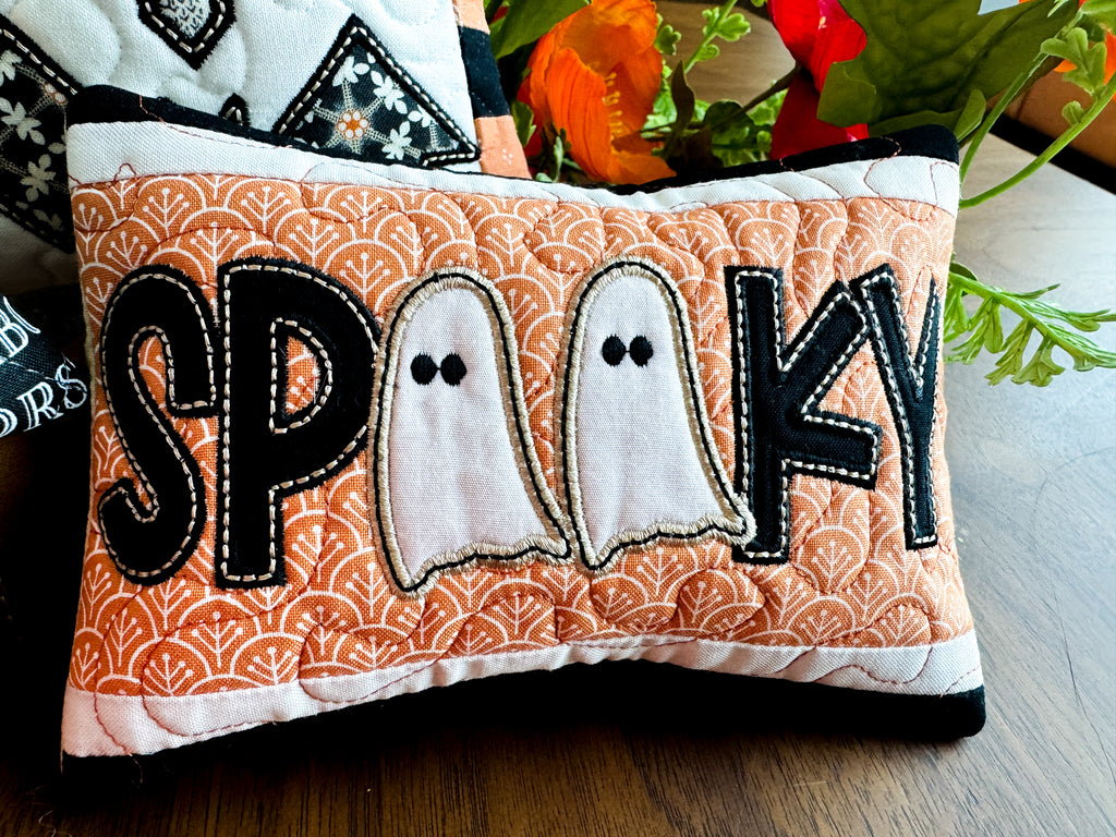 Decorative pillow with 'SPooky' text and ghost designs on an orange patterned background.