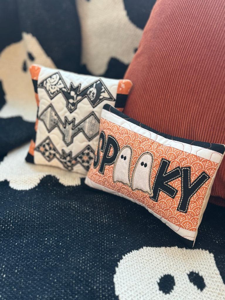 Two Halloween-themed pillows with bat and ghost designs on a textured fabric background.