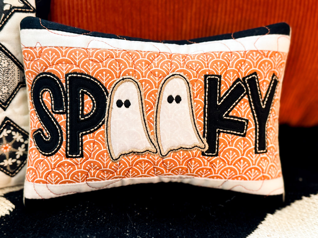 Decorative pillow with 'SPooky' text and ghost designs on an orange patterned background.