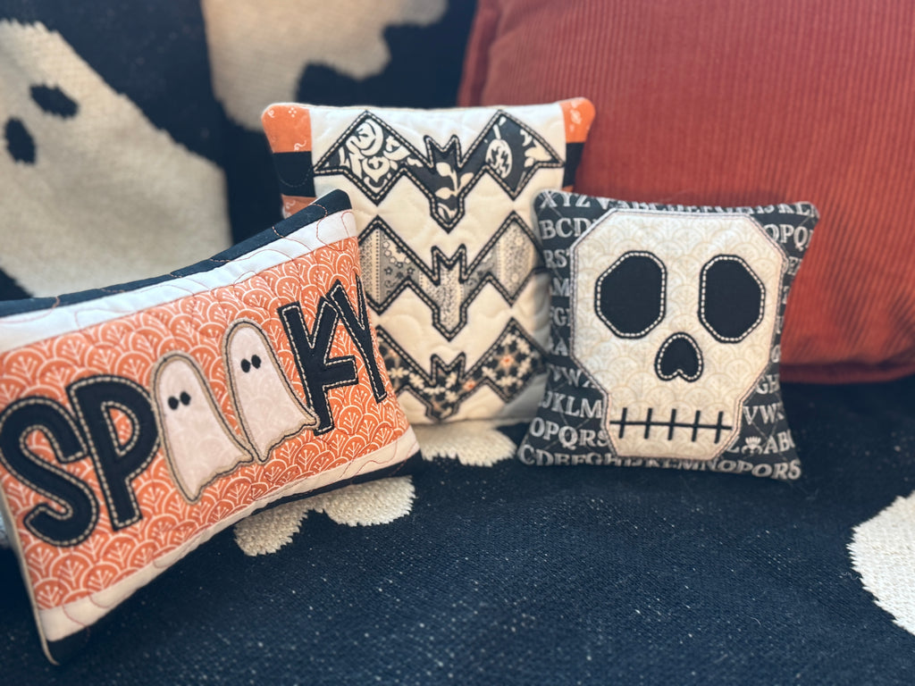 Three Halloween-themed pillows with ghost, bat, and skull designs on a dark surface.