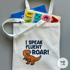 White tote bag with dinosaur graphic and text, placed on a gray surface with alphabet blocks.