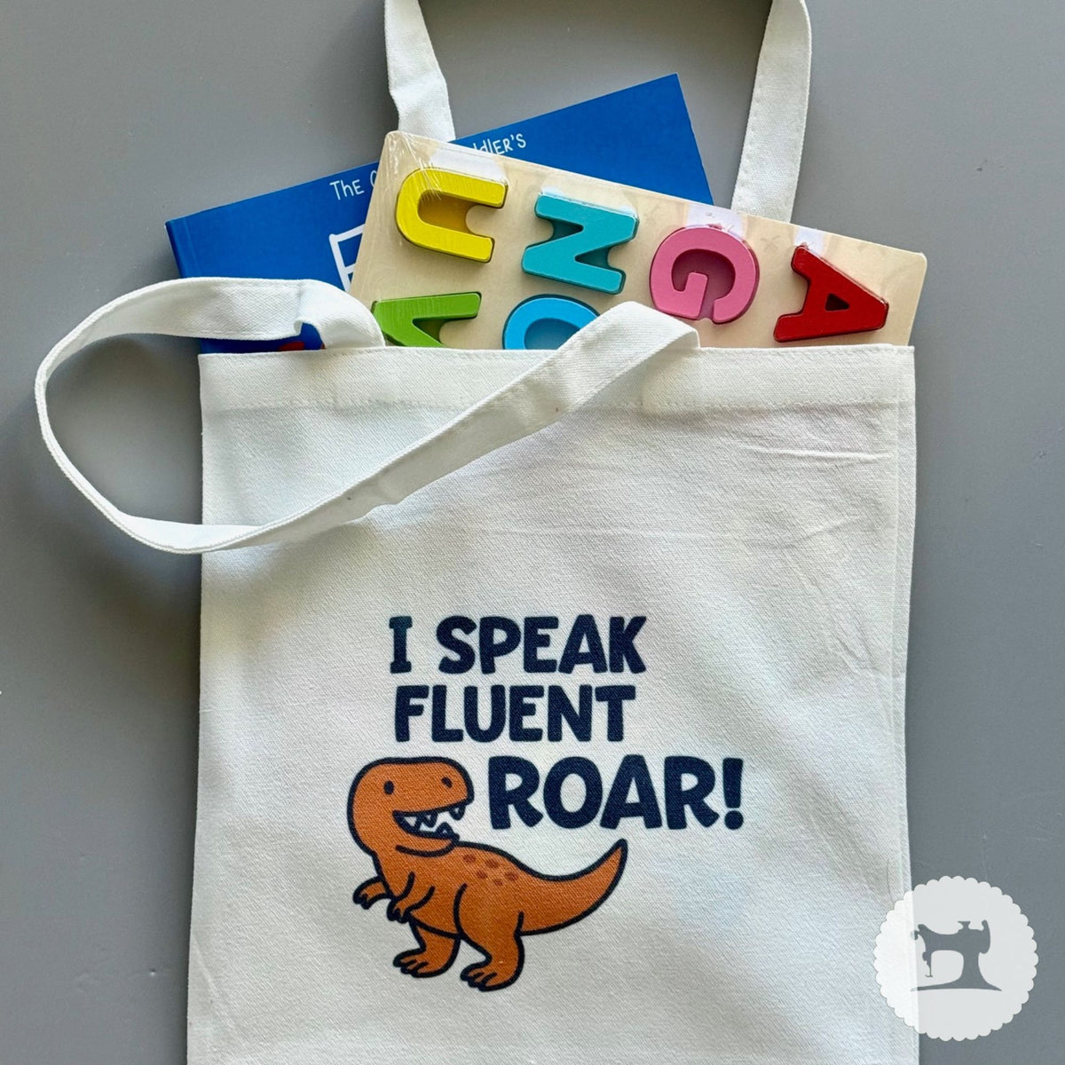White tote bag with dinosaur graphic and text, placed on a gray surface with alphabet blocks.