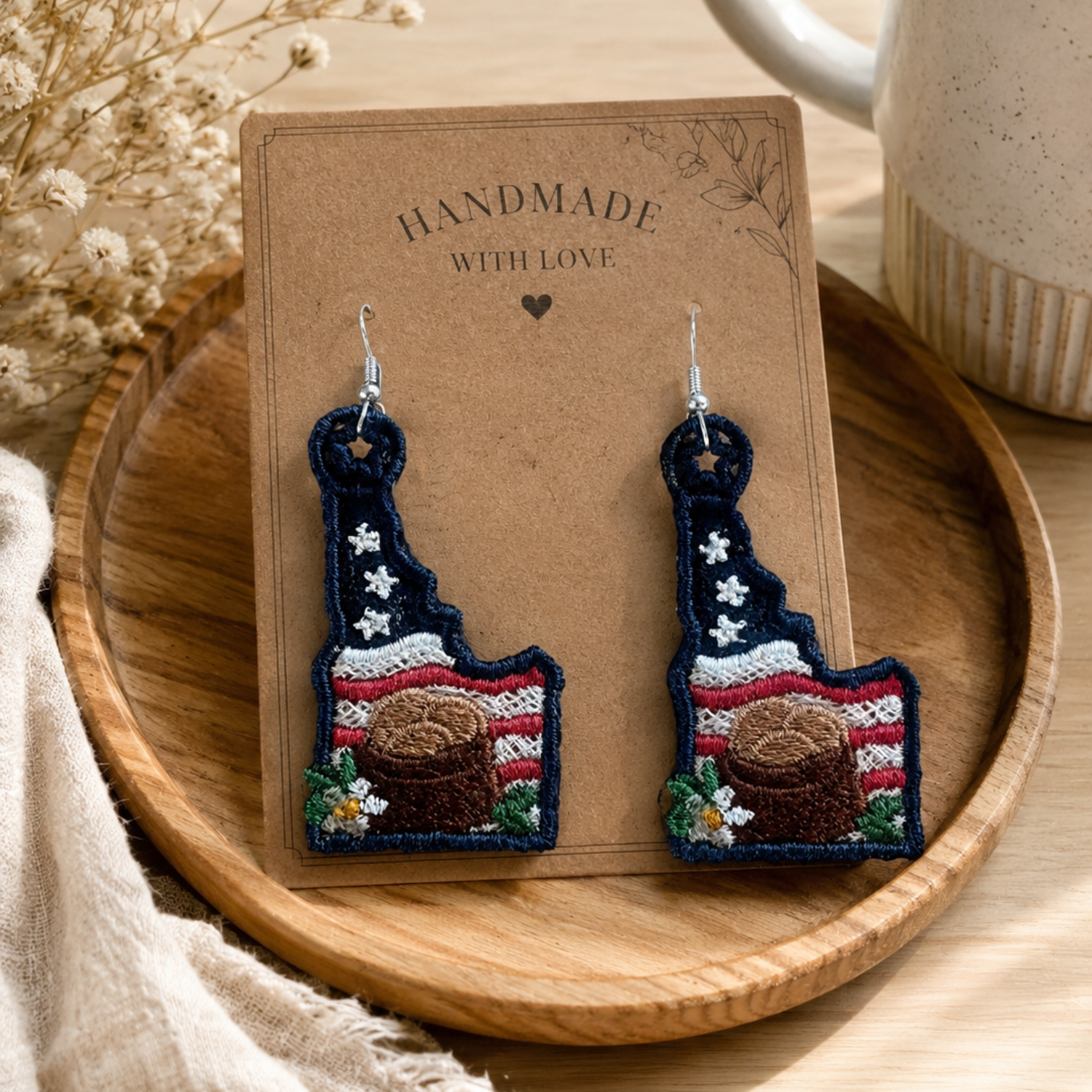 Handmade earrings on a wooden display card with a cup and dried flowers in the background.