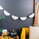 A Halloween-themed bat banner with black spiderweb designs hangs on a dark gray wall in a styled room setting with a yellow armchair, decorative pumpkins, and a plant.