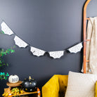 A Halloween-themed spider web banner garland hanging on a dark charcoal wall above a mustard yellow armchair and a small wooden side table with pumpkins and fall decor.
