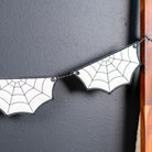 A Halloween banner garland made of bat-shaped white felt pieces with black spiderweb patterns, hanging against a dark gray wall next to a wooden ladder and beige curtain.
