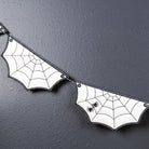 White bat-shaped banner flags with black spiderweb patterns and small black spider decorations, strung together against a dark textured wall with a small green plant branch in the corner.