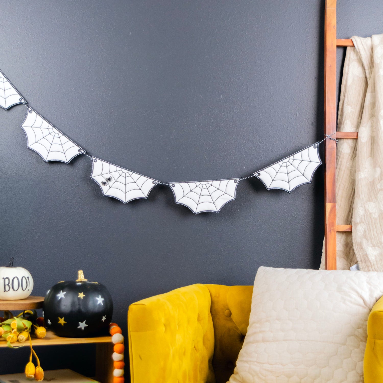 A styled interior corner with a mustard yellow armchair, white pillow, dark charcoal wall, spiderweb banner, small wooden side table with Halloween decor, and a wooden ladder leaning against the wall.