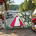A felt camping-themed tissue box cover sits on a wooden table outdoors, with a lantern, mug, marshmallows, and campsite background visible. The cover has a gray border, a green-and-white plaid background, black embroidered text reading 'NO S'MORE SNIFFLES,' a pink tent motif, and a black geometric star design.