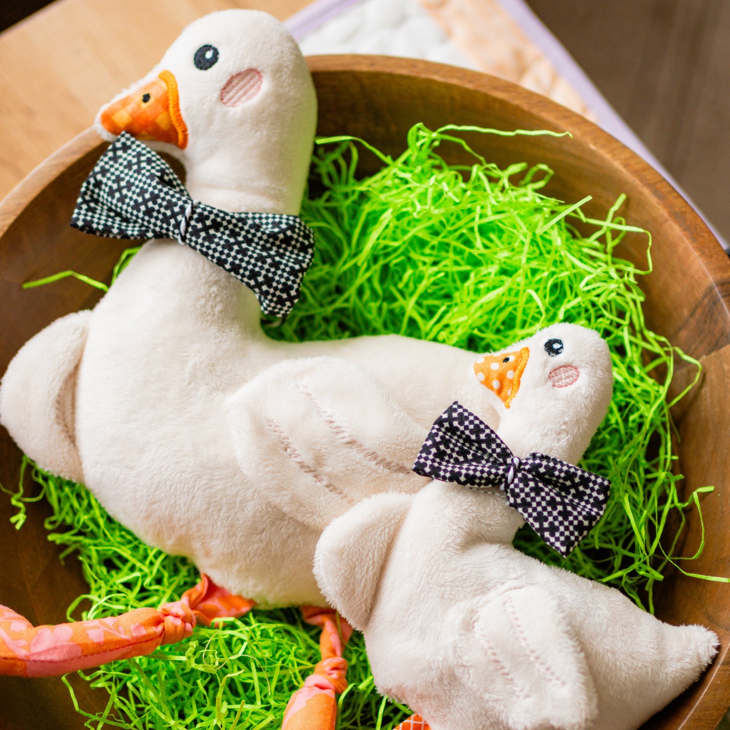 Two white plush ducks with black bow ties in a wooden bowl with green grass and carrots.