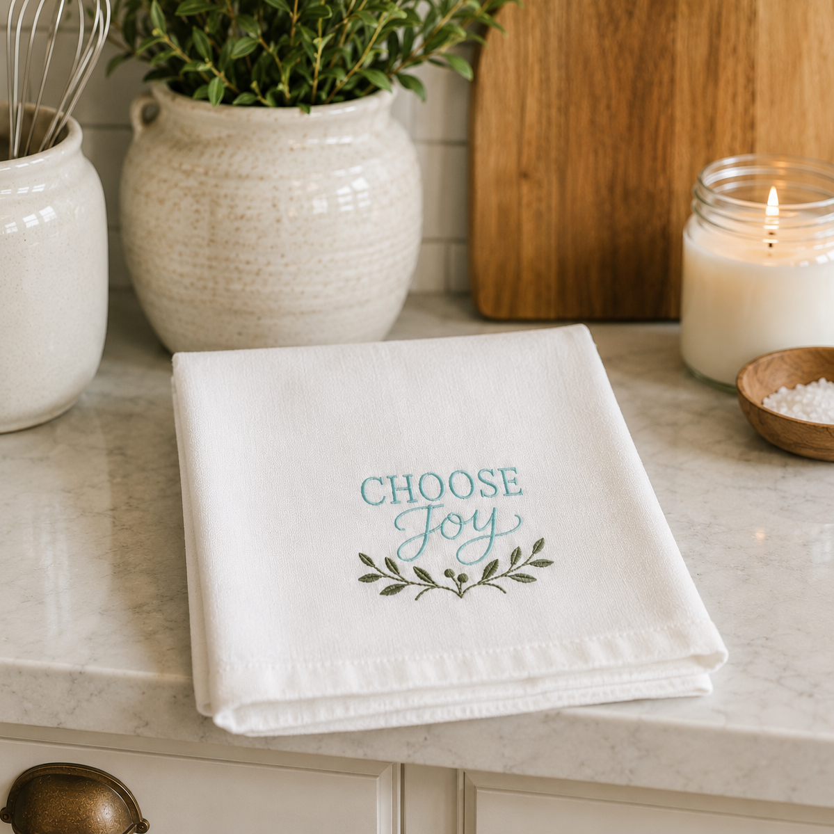 A folded white kitchen towel with the words 'CHOOSE Joy' in teal and a small leafy branch design below, displayed on a marble countertop in a styled kitchen setting.