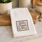 A folded white kitchen towel with 'Hello Friend' embroidered in purple and gold, resting on a wooden cutting board on a marble countertop.