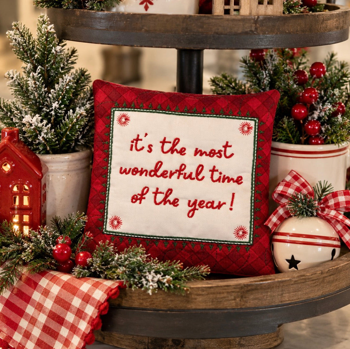 A square red holiday throw pillow with a white center panel that reads 'it's the most wonderful time of the year!' in red script, styled on a rustic wooden tiered stand with Christmas greenery, red berries, a red gingham cloth, a small red house lantern, and a white ornament.