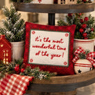 A square red holiday throw pillow with a white center panel that reads 'it's the most wonderful time of the year!' in red script, styled on a rustic wooden tiered stand with Christmas greenery, red berries, a red gingham cloth, a small red house lantern, and a white ornament.