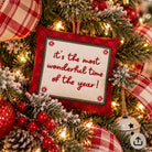 A square red Christmas throw pillow with the phrase 'it's the most wonderful time of the year!' in red embroidery, displayed on a decorated Christmas tree with lights, ornaments, and plaid ribbons.