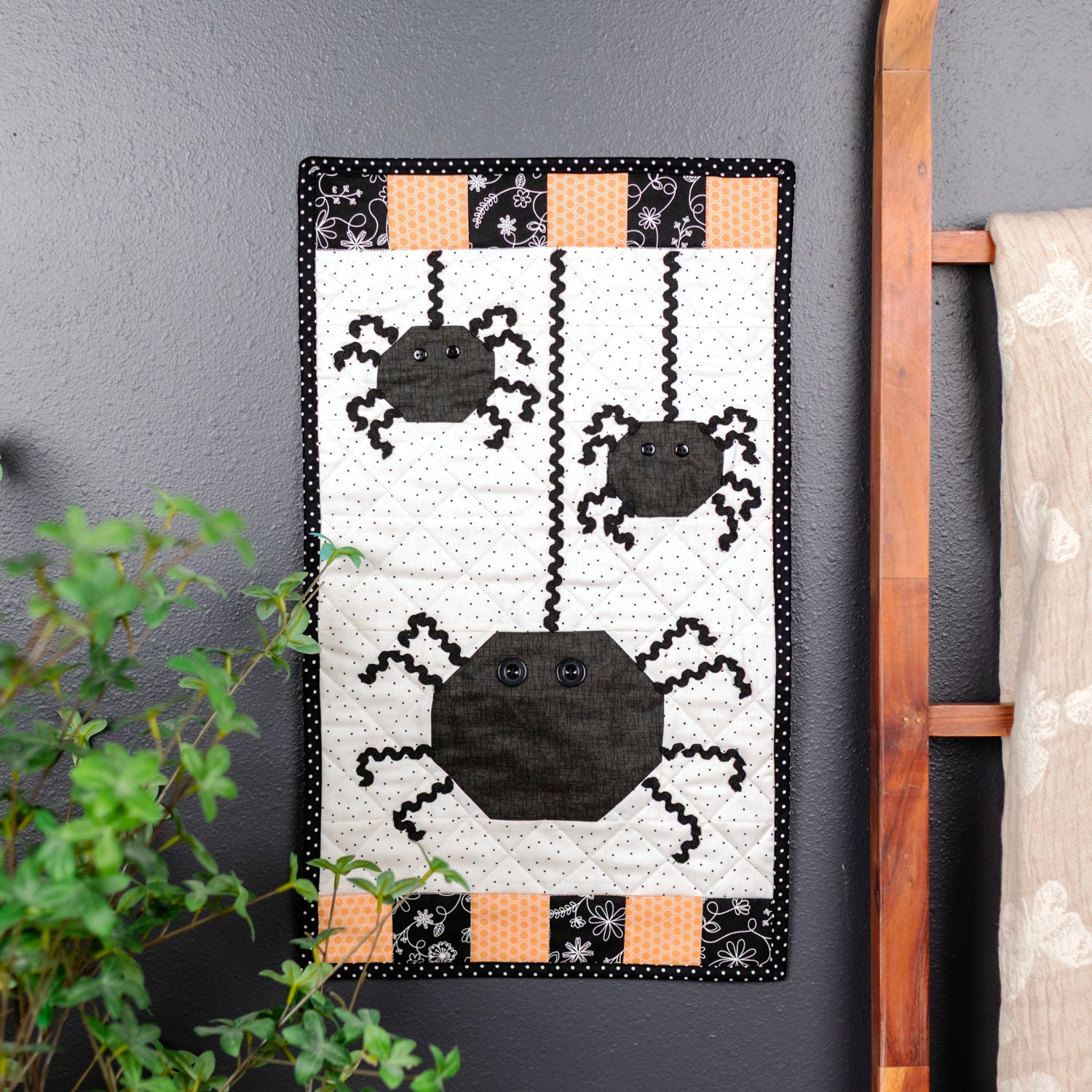 A Halloween-themed wall hanging quilt or tapestry featuring three black spiders on a white background with orange and black checkered borders, displayed on a dark gray wall next to a wooden ladder holding a patterned blanket.