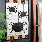 A handmade quilted wall hanging featuring three black spider motifs on an off-white background with orange and black patterned borders, displayed next to a wooden ladder holding a cream-colored fabric throw against a dark gray wall.