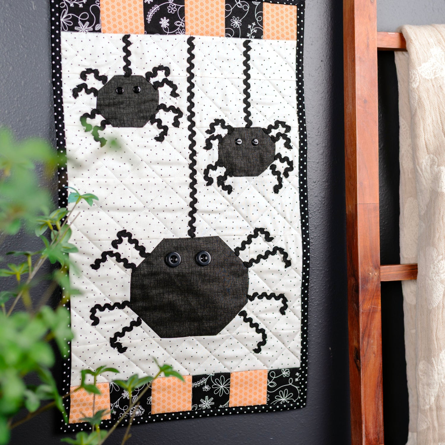 A handmade quilted wall hanging featuring three black spider motifs on an off-white background with orange and black patterned borders, displayed next to a wooden ladder holding a cream-colored fabric throw against a dark gray wall.