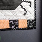 A close-up of a quilted textile piece with geometric and floral fabric patterns in black, white, and orange, shown hanging or resting against a dark gray wall next to a wooden post.