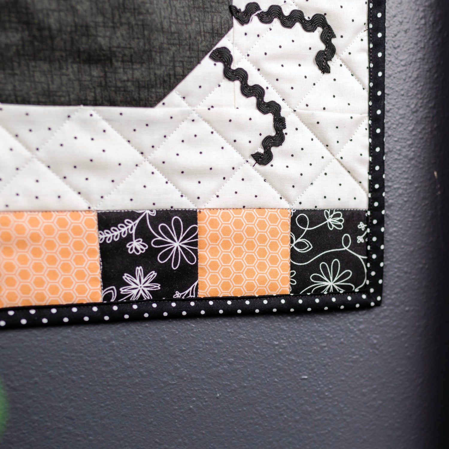 A close-up of a quilted textile piece with geometric and floral fabric patterns in black, white, and orange, shown hanging or resting against a dark gray wall next to a wooden post.