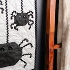 A close-up of a Halloween-themed quilted wall hanging or tapestry with black spider appliqués on a white dotted background, orange patterned borders, and a black polka-dot edge, shown hanging next to a wooden ladder and a cream textile.