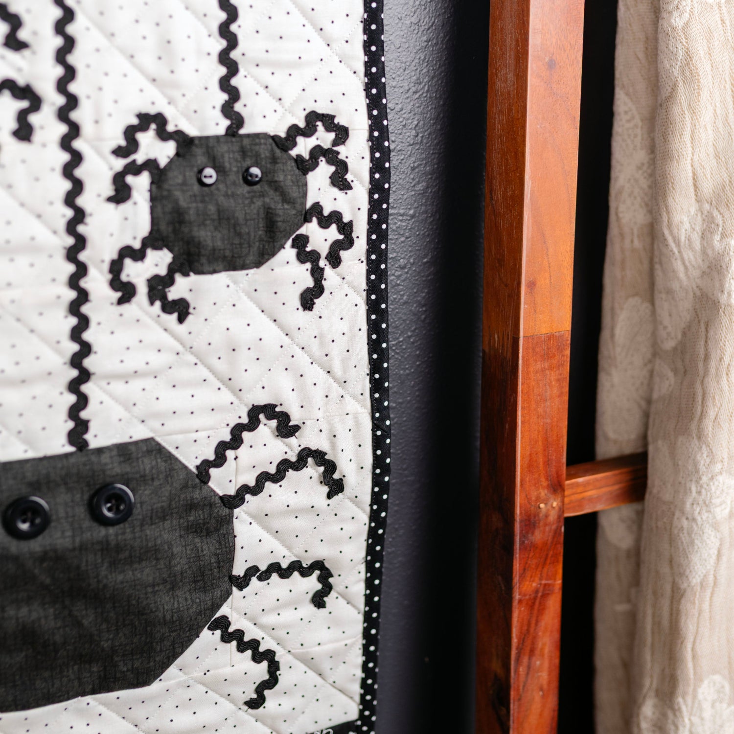 A close-up of a Halloween-themed quilted wall hanging or tapestry with black spider appliqués on a white dotted background, orange patterned borders, and a black polka-dot edge, shown hanging next to a wooden ladder and a cream textile.