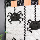 A close-up of a Halloween-themed quilted textile featuring two black spider appliqués on a white dotted background, with black-and-white polka dot trim, orange and black patterned fabric accents, and a dark gray wall behind it.