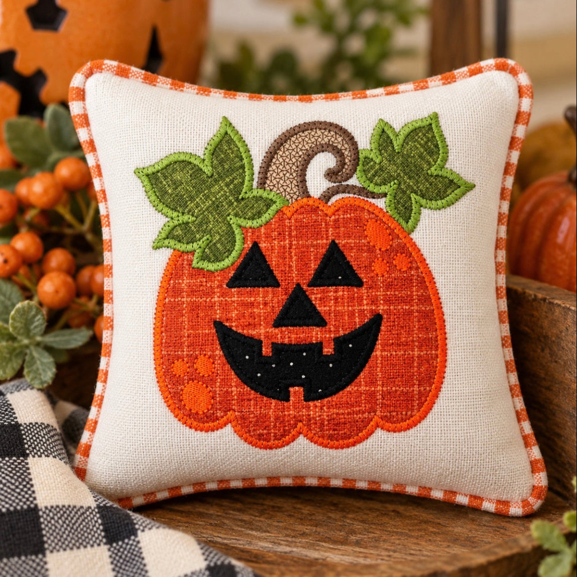 A square decorative throw pillow with a stitched jack-o'-lantern design, featuring an orange pumpkin face with green leaves and a brown stem on a cream fabric background with an orange-and-white checkered border. The pillow is styled on a wooden surface with autumn decorations in the background.