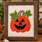 A framed autumn-themed needlepoint or embroidery featuring a smiling orange jack-o'-lantern with green leaves and a brown stem on a beige fabric background, set in a dark wood frame with seasonal decor around it.