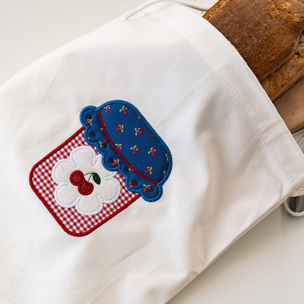 White towel with a decorative patch featuring a blue jar, red checkered base, and white flower with a cherry on top.