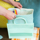 Person holding a teal basket with stationery items against a blurred indoor background