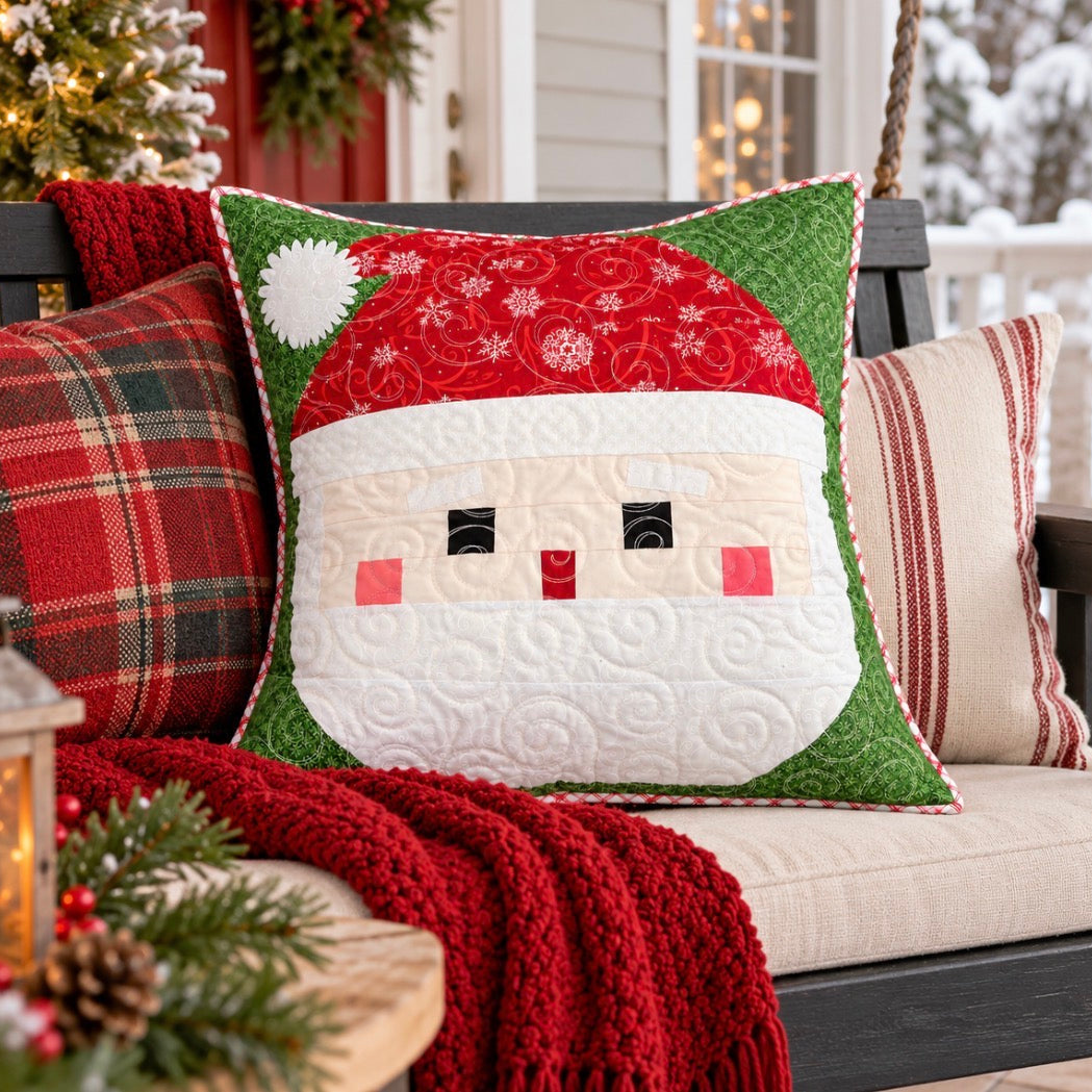 A festive Christmas-themed quilted throw pillow featuring a smiling Santa Claus face, styled on a porch swing with a red knit blanket, a red plaid pillow, and a striped pillow against a snowy outdoor background.