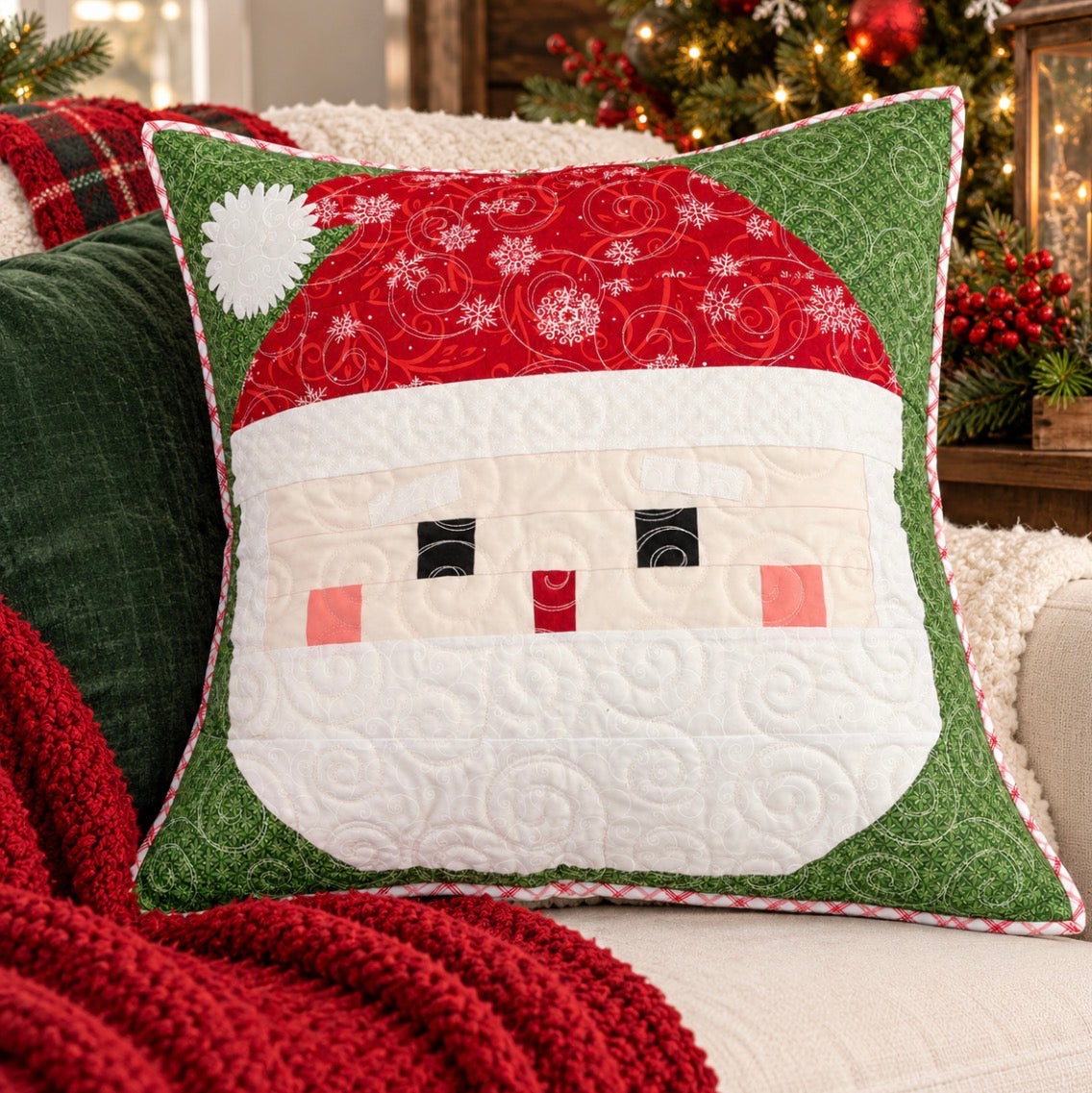 A square Christmas-themed quilted throw pillow shaped like Santa's face, styled on a cozy white sofa with a red knit blanket and a blurred festive Christmas tree in the background.