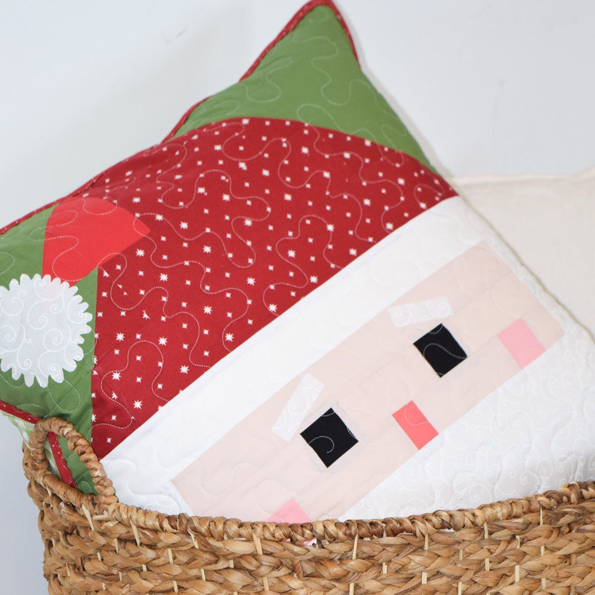 Decorative pillow with a Santa face design in a woven basket against a white wall.