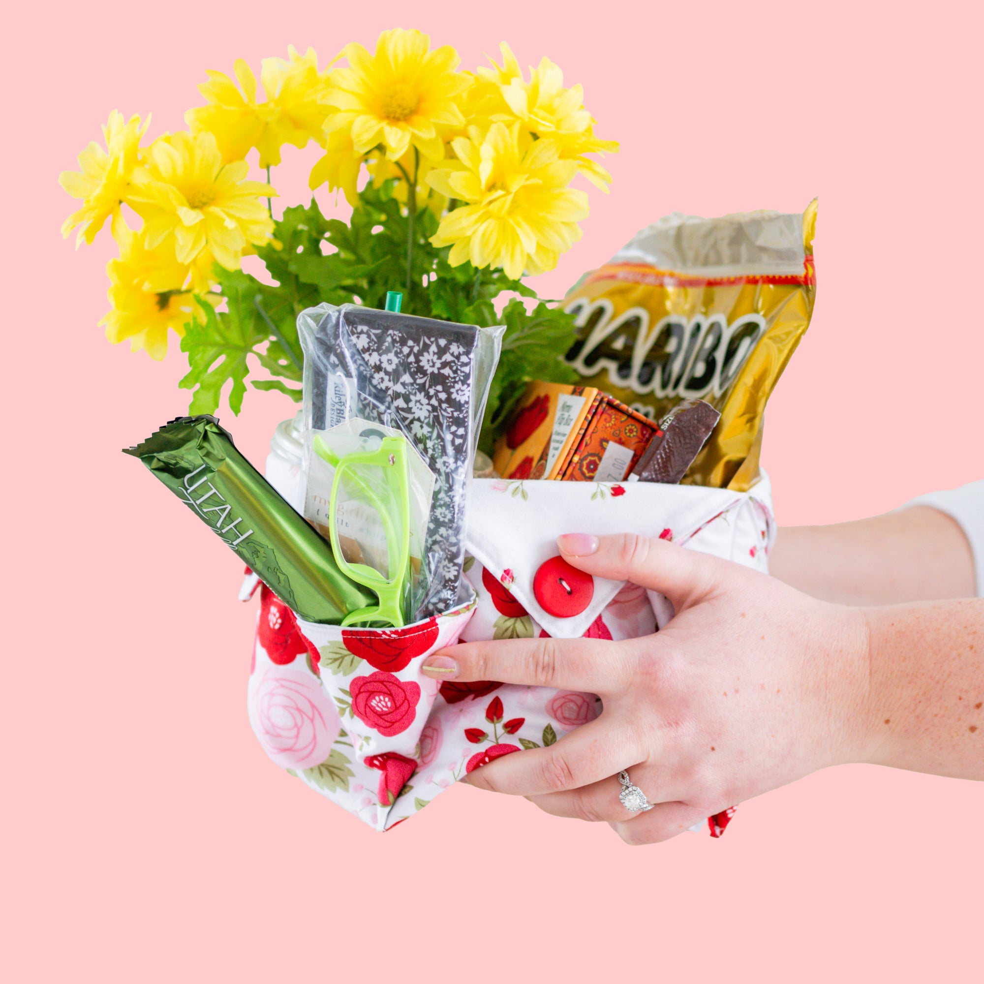 Hand holding a colorful fabric basket with snacks and flowers against a pink background