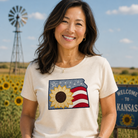 Woman wearing a t-shirt with a sunflower and American flag design, standing in a sunflower field with a windmill and 'Welcome to Kansas' sign in the background.